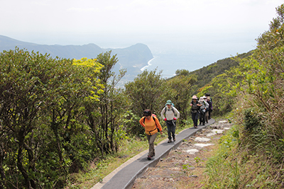 後ろに海を見ながら、登山道の階段横のコンクリート道を登っている写真